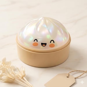 May include: A beige and iridescent white dumpling-shaped night light with a smiling face. The night light has a round base and a textured, dome-shaped top. The background includes a sprig of dried flowers and a blank tag.