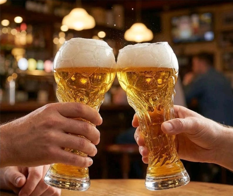 May include: Two clear glass beer mugs with a textured exterior resembling the hexagonal pattern of a soccer ball. Each glass is filled with golden beer topped with a thick white foam head, held up in a celebratory toast against a blurred background.
