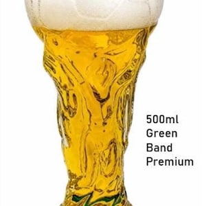 May include: A clear glass beer mug shaped like a trophy, filled with golden liquid and white foam. The base features a green band. Text on the right reads 500ml Green Band Premium. The glass has a textured surface resembling a soccer ball pattern.