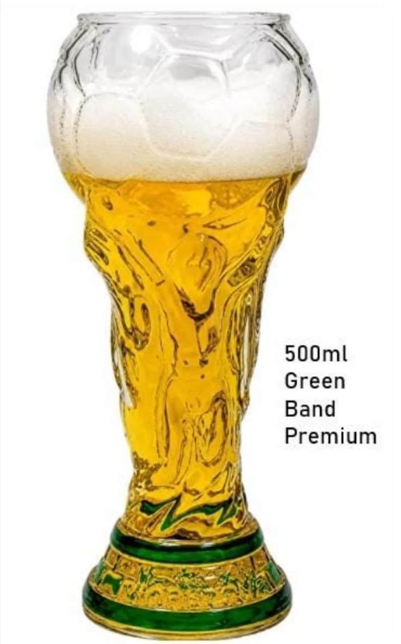May include: A clear glass beer mug shaped like a trophy, filled with golden liquid and white foam. The base features a green band. Text on the right reads 500ml Green Band Premium. The glass has a textured surface resembling a soccer ball pattern.