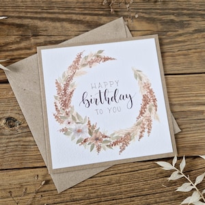 May include: A square greeting card with a brown kraft paper envelope featuring a watercolour wreath of dried flowers and grasses in shades of beige, brown, and muted green. The text "HAPPY birthday TO YOU" is printed in the centre in black ink.
