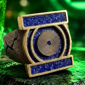 May include: A metallic ring featuring a rectangular top with blue stone inlays and a central circular emblem with concentric rings. The band has a textured, weathered finish, and the design is inspired by the Green Lantern Corps symbol.