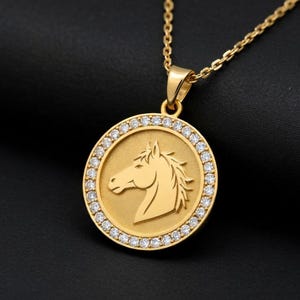 Puede incluir: Un collar con colgante dorado con un diseño de cabeza de caballo. El colgante circular tiene un borde de pequeñas piedras preciosas transparentes. La silueta del caballo está en el centro. La cadena y el colgante son de metal dorado.