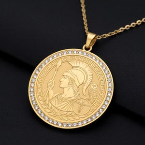 May include: A gold-colored pendant necklace featuring a circular medallion with a detailed relief of a figure in a helmet holding a spear. The medallion is encircled by small, clear gemstones and a decorative border. The necklace chain is also gold-colored.