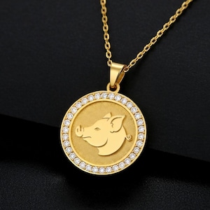 Puede incluir: Collar con colgante dorado con un colgante circular con la silueta de un cerdo. El colgante está rodeado de pequeñas piedras preciosas transparentes. La cadena y el colgante son dorados, creando un diseño elegante.