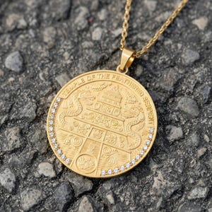 May include: Gold-colored pendant necklace with a round charm. The charm features the text "TEMPLE OF THE FEATHERED SERPENT" and an engraved design with two serpents and a central structure. Small, clear stones encircle the charm.