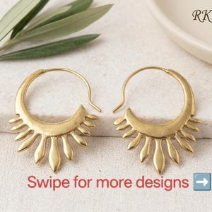Può includere: Un paio di orecchini a cerchio color oro con un design a raggiera che presenta forme a petalo lungo il bordo inferiore. Il testo RK E045 è visibile nell'angolo in alto a destra e il testo Swipe for more designs con una freccia in basso.