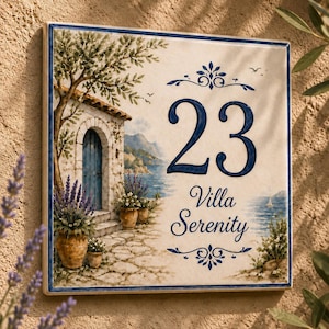Pode incluir: Uma placa de casa quadrada de cerâmica com uma cena mediterrânea pintada, apresentando uma villa de pedra, porta azul e vasos de lavanda. O número 23 está em azul escuro acima do texto Villa Serenity em estilo de escrita cursiva.