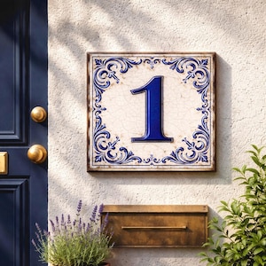 Peut inclure: Plaque de numéro de maison carrée en céramique avec le chiffre « 1 » bleu foncé. Le chiffre est centré sur un fond crème craquelé, encadré de volutes bleues. La plaque est fixée sur un mur blanc texturé, avec une boîte aux lettres en laiton en dessous.