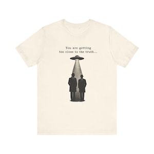 Puede incluir: Camiseta color crema con una ilustración en blanco y negro de dos hombres con trajes y sombreros, de pie debajo de un OVNI. El texto "You are getting too close to the truth..." está impreso encima del OVNI.