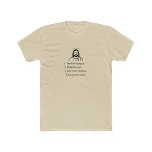 以下が含まれることがあります： 薄いタン色のTシャツ。ハローのある人物のグラフィックと「1. 飢えた人に食べ物を、2. 貧しい人を助け、3. 敵を愛しなさい。私はとても明確でした。」というテキストが特徴です。このシャツは柔らかい素材で作られています。