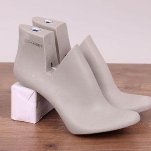 Puede incluir: Dos hormas de zapatos grises con tacones altos, apoyadas sobre un bloque envuelto en papel blanco, contra un fondo claro. Las hormas están hechas de un material sólido y están diseñadas para dar forma al calzado. El texto "2CO-HOBBY" es visible.