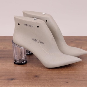 May include: A pair of light grey shoe lasts with clear acrylic heels. The lasts have pointed toes and are marked with the text "B131121" and "25-06-2021". The heels are approximately 7.6 cm tall.