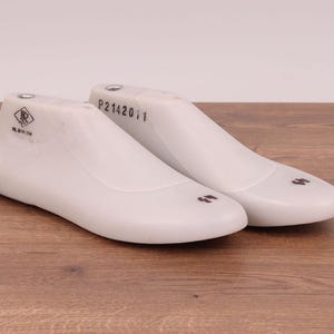 Puede incluir: Dos hormas de zapatos blancas, utilizadas en la fabricación de calzado, se colocan sobre una superficie de madera. Cada horma tiene una punta redondeada y un talón, con marcas y el número "11" visible. Una horma tiene el texto "P2142011".