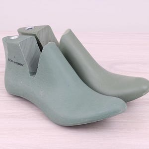 May include: Two sage green shoe lasts, one with the text "ECO-HOBBY" printed on the side. The lasts are made of a smooth material and are designed to maintain the shape of shoes. They are placed on a light wood grain surface.