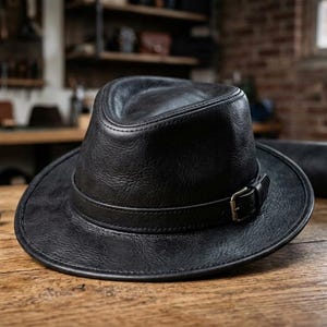 Może przedstawiać: Czarny skórzany kapelusz typu fedora z wgniecioną koroną i szerokim rondem. Pasujący czarny skórzany pasek owija się wokół podstawy korony, zabezpieczony małą metalową klamrą z boku. Kapelusz posiada widoczną ziarnistą fakturę materiału.