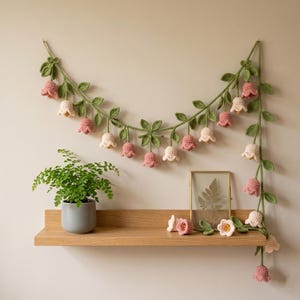 May include: A handmade crochet garland featuring pink and cream-colored bell-shaped flowers and green leaves. The garland is displayed on a wall, with a wooden shelf below holding a potted plant and decorative items. The garland is approximately 60 inches long.