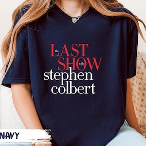 May include: Navy blue t-shirt with the text "the LAST SHOW with stephen colbert" in red and white lettering. The shirt is a crew neck style.