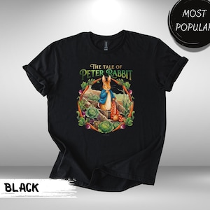 May include: Black t-shirt featuring a colorful illustration from "The Tale of Peter Rabbit." The design includes Peter Rabbit in a blue jacket, surrounded by vegetables and the text "The Tale of Peter Rabbit." A "MOST POPULAR" badge is visible.