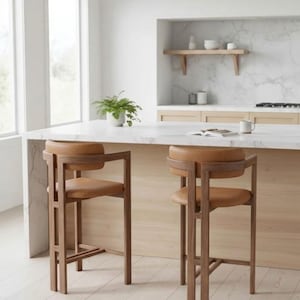 Könnte beinhalten: Zwei Barhocker aus Holz mit braunen Ledersitzen und Rückenlehnen. Die Hocker haben ein modernes Design mit einem rechteckigen Rahmen und stehen in einer Küche mit einer Marmorarbeitsplatte.