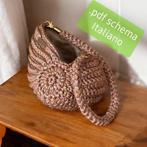 May include: A handcrafted, iridescent pink crochet bag with a braided handle and gold-toned hardware. The bag has a unique, textured design and a neutral-colored interior. The image includes a green rectangle with the text ".pdf schema Italiano."