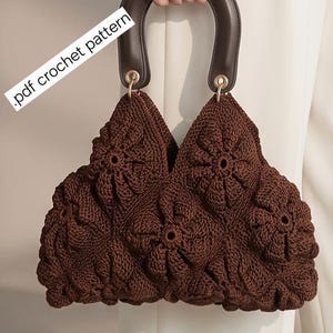 May include: A dark brown crochet handbag featuring a textured floral motif pattern throughout the body. The bag includes a solid dark brown arched handle attached with gold-tone hardware. The text .pdf crochet pattern is visible on the side.