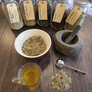 K&ouml;nnte beinhalten: Eine Auswahl an getrockneten Kr&auml;utern und Zutaten f&uuml;r Kr&auml;utermittel. Enth&auml;lt Gl&auml;ser mit den Aufschriften Ashwagandha Root, Ginger Root, Lemon Balm Leaf, Rosehips und Oatstraw. Eine Tasse Tee, ein M&ouml;rser und St&ouml;&szlig;el sowie ein kleiner L&ouml;ffel sind ebenfalls vorhanden.