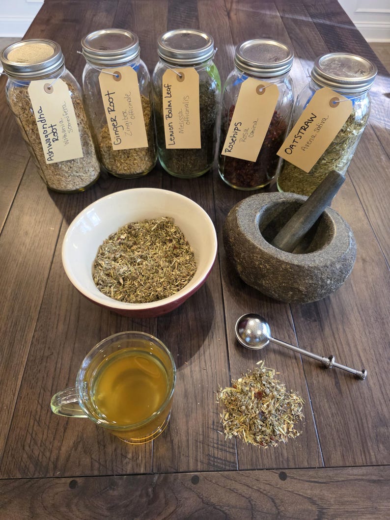 K&ouml;nnte beinhalten: Eine Auswahl an getrockneten Kr&auml;utern und Zutaten f&uuml;r Kr&auml;utermittel. Enth&auml;lt Gl&auml;ser mit den Aufschriften Ashwagandha Root, Ginger Root, Lemon Balm Leaf, Rosehips und Oatstraw. Eine Tasse Tee, ein M&ouml;rser und St&ouml;&szlig;el sowie ein kleiner L&ouml;ffel sind ebenfalls vorhanden.