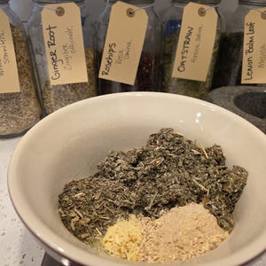 K&ouml;nnte beinhalten: Eine wei&szlig;e Keramiksch&uuml;ssel gef&uuml;llt mit getrockneten Kr&auml;utern und Gew&uuml;rzen. Die Sch&uuml;ssel enth&auml;lt eine Mischung aus gr&uuml;nen, braunen und beigen Zutaten. Im Hintergrund zeigen Gl&auml;ser mit handgeschriebenen Etiketten die Namen verschiedener Kr&auml;uter, darunter "Ashwagandha Root" und "Ginger Root."