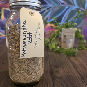 May include: A clear glass jar filled with dried Ashwagandha Root. A cream-colored tag is attached to the jar with the words "Ashwagandha Root" and "Withania Somnifera" written on it. The jar has a silver lid. The background is a mix of green and blue.