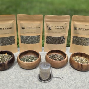 May include: Four tan paper pouches labeled First Light, Anchor and Root, Crimson Harmony, and Iron Twilight, each containing loose tea. Four small round wooden bowls filled with tea leaves sit in front, with a metal tea infuser in the center.