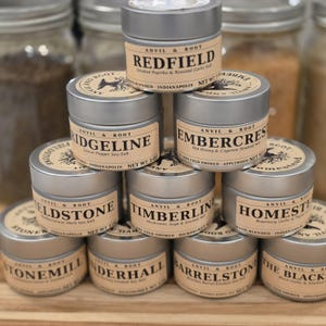 May include: A collection of small, silver metal tins of gourmet spices, stacked on a wooden surface. Each tin has a label with the brand name "Anvil & Root" and a different spice name, such as "Redfield" and "Embercres". The background includes glass jars.