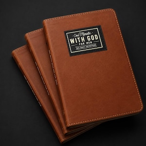 May include: Three brown leather-bound books stacked together. Each cover features a black rectangular label with gold text that reads One Minute WITH GOD FOR MEN 365 DAILY DEVOTIONS. The books have stitched edges and a smooth, matte finish.