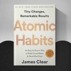 May include: A book titled Atomic Habits by James Clear with a white cover featuring gold speckled text. The cover includes the subtitle Tiny Changes, Remarkable Results, An Easy & Proven Way to Build Good Habits & Break Bad Ones, and a gold seal.
