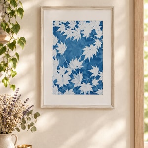 Pode incluir: Uma impressão de arte de parede retangular numa moldura de madeira clara com um design botânico estilo cianotipia. A obra exibe silhuetas brancas de folhas de ácer japonês sobrepostas num fundo azul profundo com textura de aguarela.