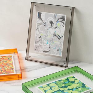 Può includere: Una cornice in acrilico trasparente con bordo grigio, che mostra una stampa astratta con vortici grigi, neri e blu. Sono visibili altre cornici arancioni e verdi, ciascuna con arte astratta colorata. Le cornici sono su una superficie bianca.