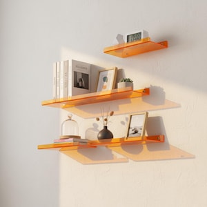 Puede incluir: Tres estantes flotantes de acrílico naranja montados en una pared blanca. Los estantes exhiben libros, fotos enmarcadas, una pequeña planta, un jarrón decorativo y un reloj digital. Los estantes son de diferentes tamaños.
