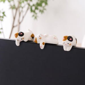 May include: Three small calico cat figurines with white, black, and orange patches. Each cat is posed in a different position, including one reaching forward, one lying on its back, and one sitting with its front paws resting over a black edge.