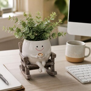 May include: A white ceramic planter shaped like a round character with a smiling face, holding a small coffee cup. The planter sits in a miniature brown wooden rocking chair and contains a green leafy plant with small, rounded foliage.