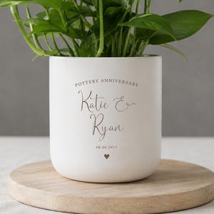 May include: A white ceramic plant pot with green foliage. The pot has rounded edges and features the text "POTTERY ANNIVERSARY" above the names "Katie & Ryan" and the date "08.06.2017" with a heart symbol.