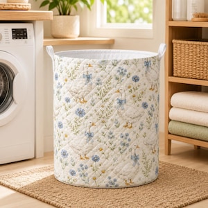 May include: A cylindrical laundry hamper featuring a quilted white fabric exterior decorated with a repeating pattern of white geese wearing blue bows, interspersed with small blue and yellow wildflowers and green stems, and two white side handles.
