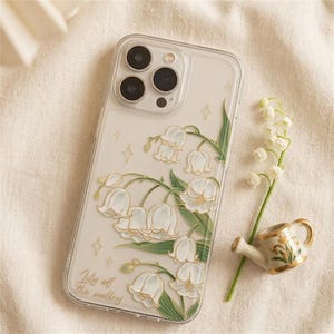 Könnte beinhalten: Transparente Handyhülle mit floralem Design mit weißen Maiglöckchenblüten und grünen Blättern. Die Hülle trägt den Text "Lily of the valley" und goldfarbene Akzente. Ein Zweig Maiglöckchen und eine Miniatur-Gießkanne liegen daneben.