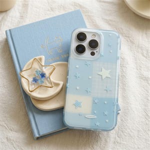 May include: A light blue phone case with a star pattern, a light blue book, and a star-shaped trinket holder. The phone case has a patchwork design with white and blue squares and small star accents.