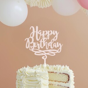 May include: A white cake with white frosting and a slice cut out, topped with a white "Happy Birthday" cake topper. White balloons and a paper fan are in the background. The cake has a layered appearance.