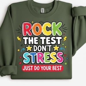 Op de afbeelding: Een olijfgroene sweatshirt met de tekst "ROCK THE TEST DON'T STRESS JUST DO YOUR BEST" in kleurrijke, dikgedrukte letters. Het ontwerp bevat sterren, bliksemschichten en harten. Een casual, motiverend kledingstuk.
