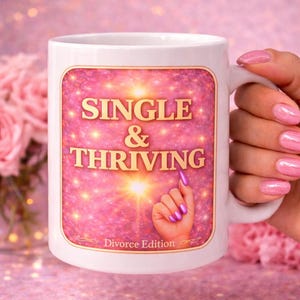 May include: White ceramic mug with the words "SINGLE & THRIVING" in gold lettering on a pink background. The mug also features a hand with purple nails and the text "Divorce Edition".