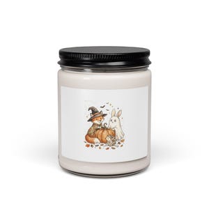 May include: A clear glass candle jar with a black lid featuring a white label. The label displays a cartoon illustration of a fox wearing a witch hat, a white ghost rabbit, and a small mummy hedgehog gathered around a pumpkin with autumn leaves.