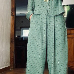 May include: A sage green two-piece set featuring a long-sleeved top with a mandarin collar, gold-toned frog closures, and wide-leg trousers. The fabric is adorned with a repeating small geometric pattern and finished with light yellow piping trim.