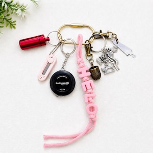May include: A collection of keychains featuring a variety of charms. Includes a red metal container, a pink cutter, a black tape measure, and charms with sewing-related themes. A pink cord spells out the word "HELLO".