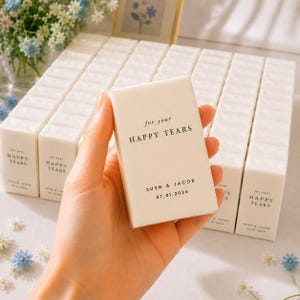 May include: An off-white box held in a hand, printed with "for your HAPPY TEARS", the names "SUSN & JACOB" and the date "07.01.2026". Other boxes with the same text are in the background, along with floral decorations.
